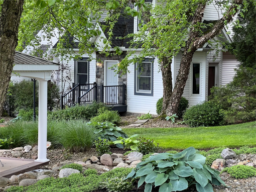 White house with stone accents, lush green landscaping, and large trees.