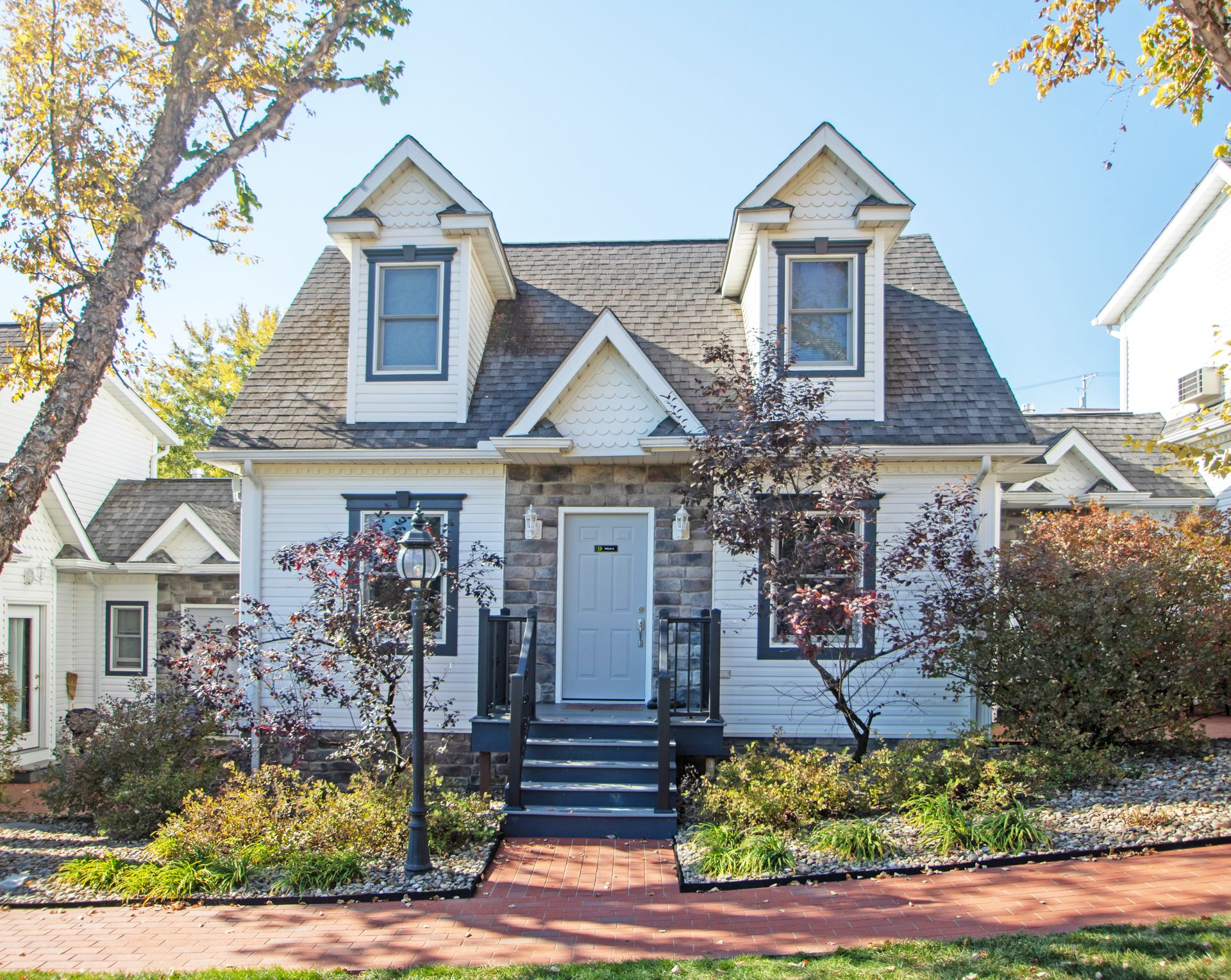 White house with stone facade, two dormers, dark trim, and a brick walkway.