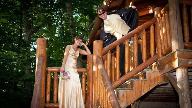 Bride in cream gown poses on wooden stairs; groom in tuxedo jacket above.