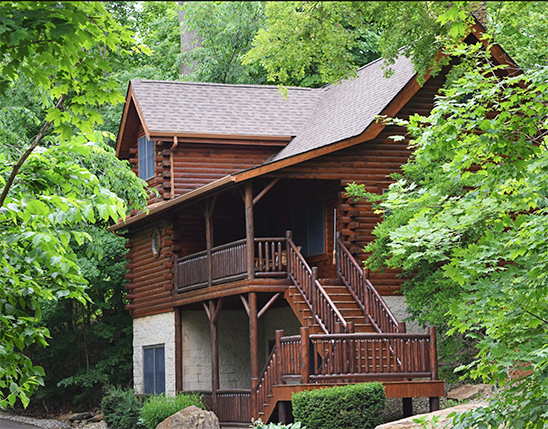 Log cabin with brown walls, wooden stairs, and a dark roof, surrounded by green foliage.