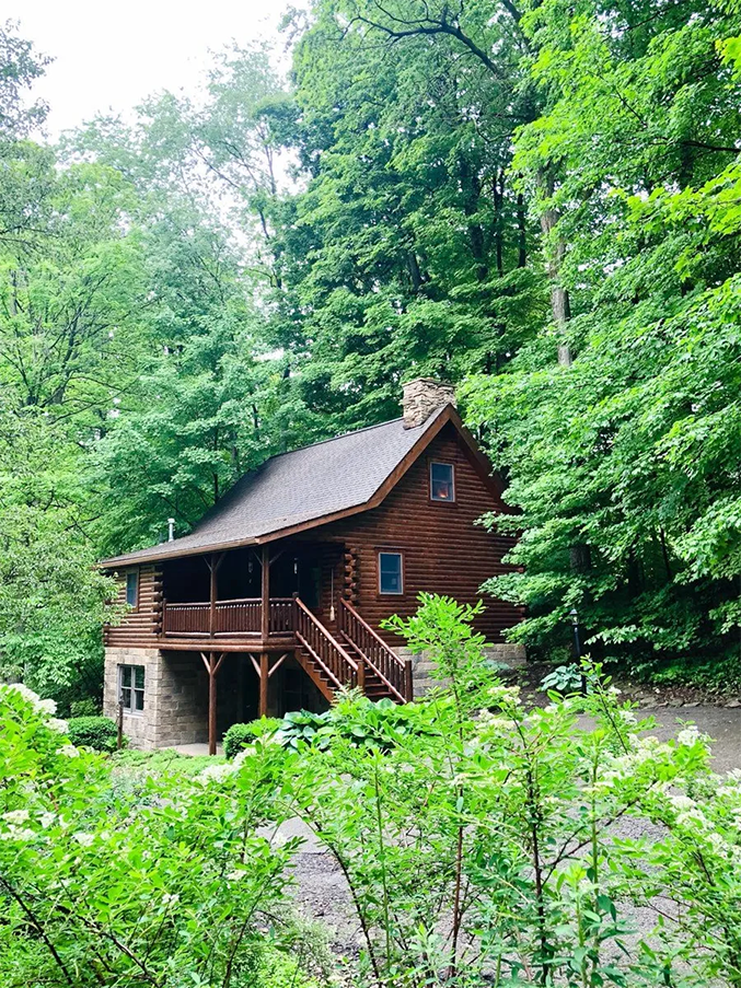 Log cabin nestled in lush green trees, with a stone chimney and wooden deck.