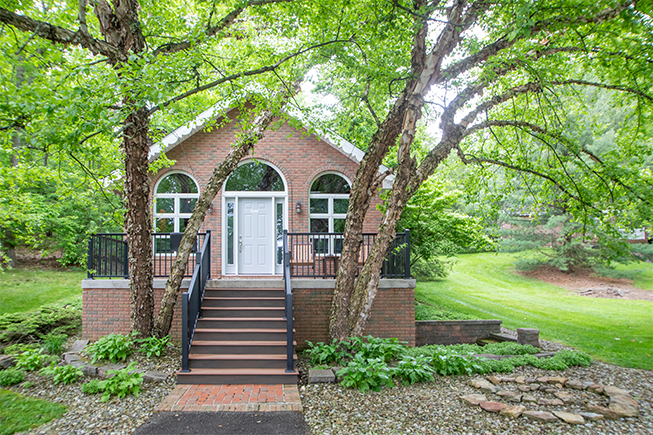 Brick building with arched windows, white door, and steps, framed by trees in a green yard.