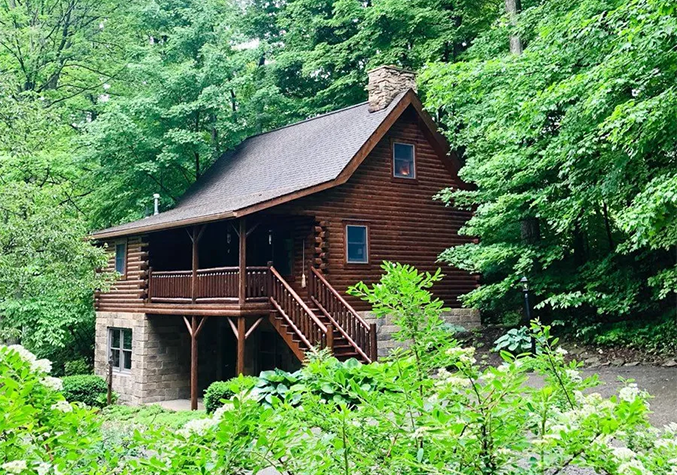 Log cabin nestled in lush green trees with a stone foundation and wooden stairs leading to a porch.