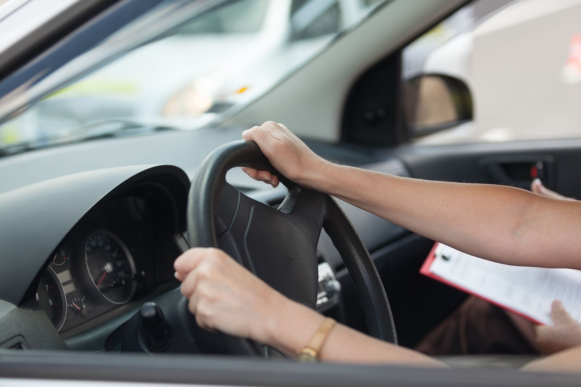 Hands gripping a steering wheel inside a car, with a clipboard in the passenger seat.