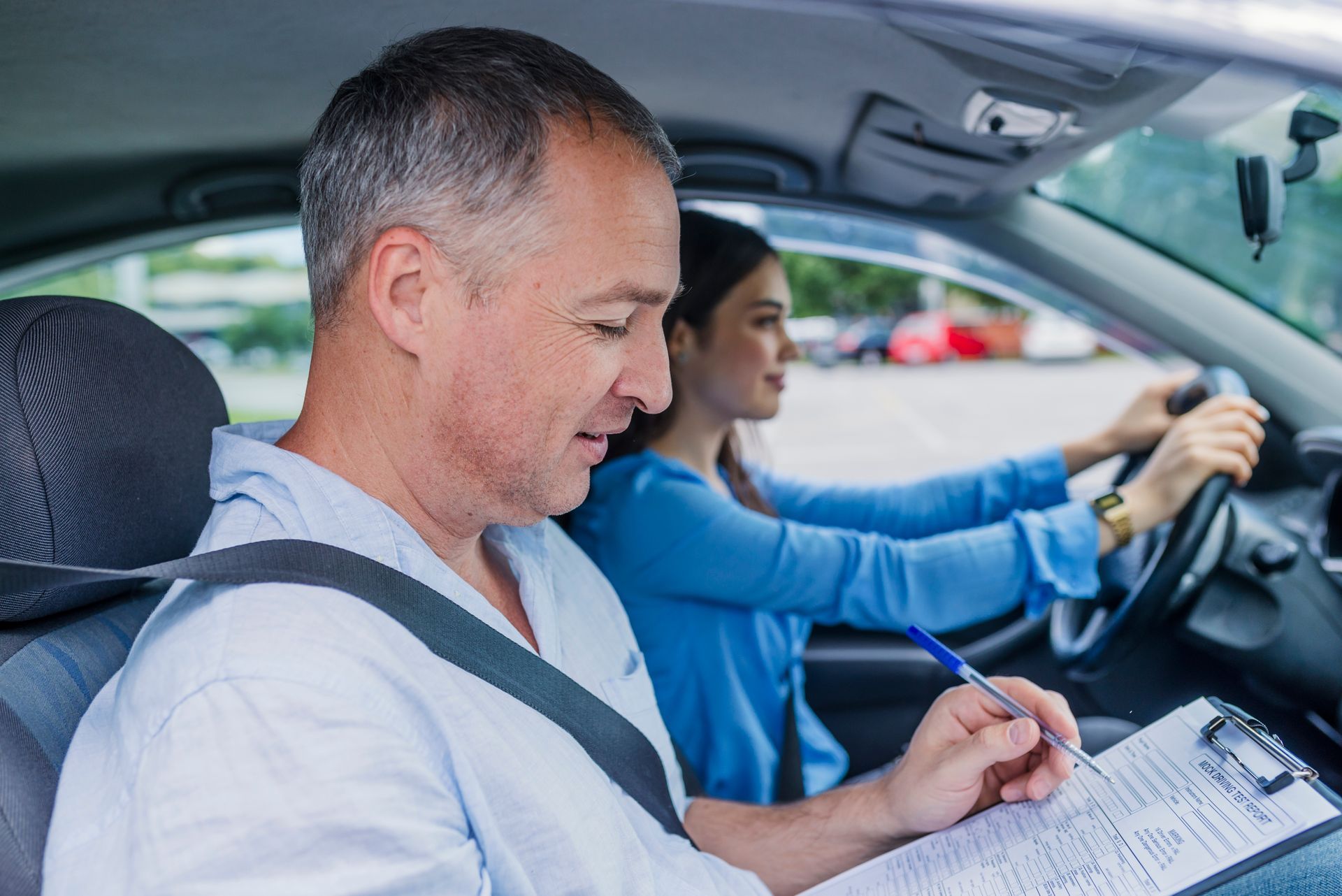 Driving instructor in car, writing on clipboard while student drives.