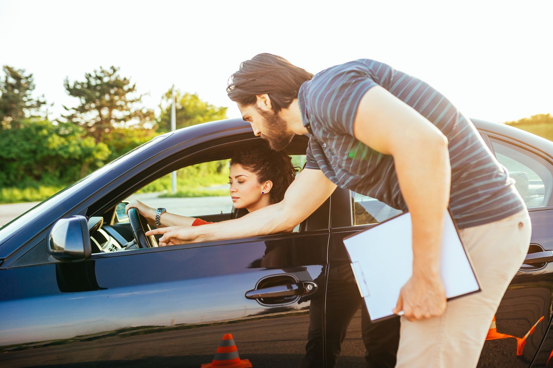 Driving instructor pointing at dashboard while teaching a person how to drive in a car, outdoors.