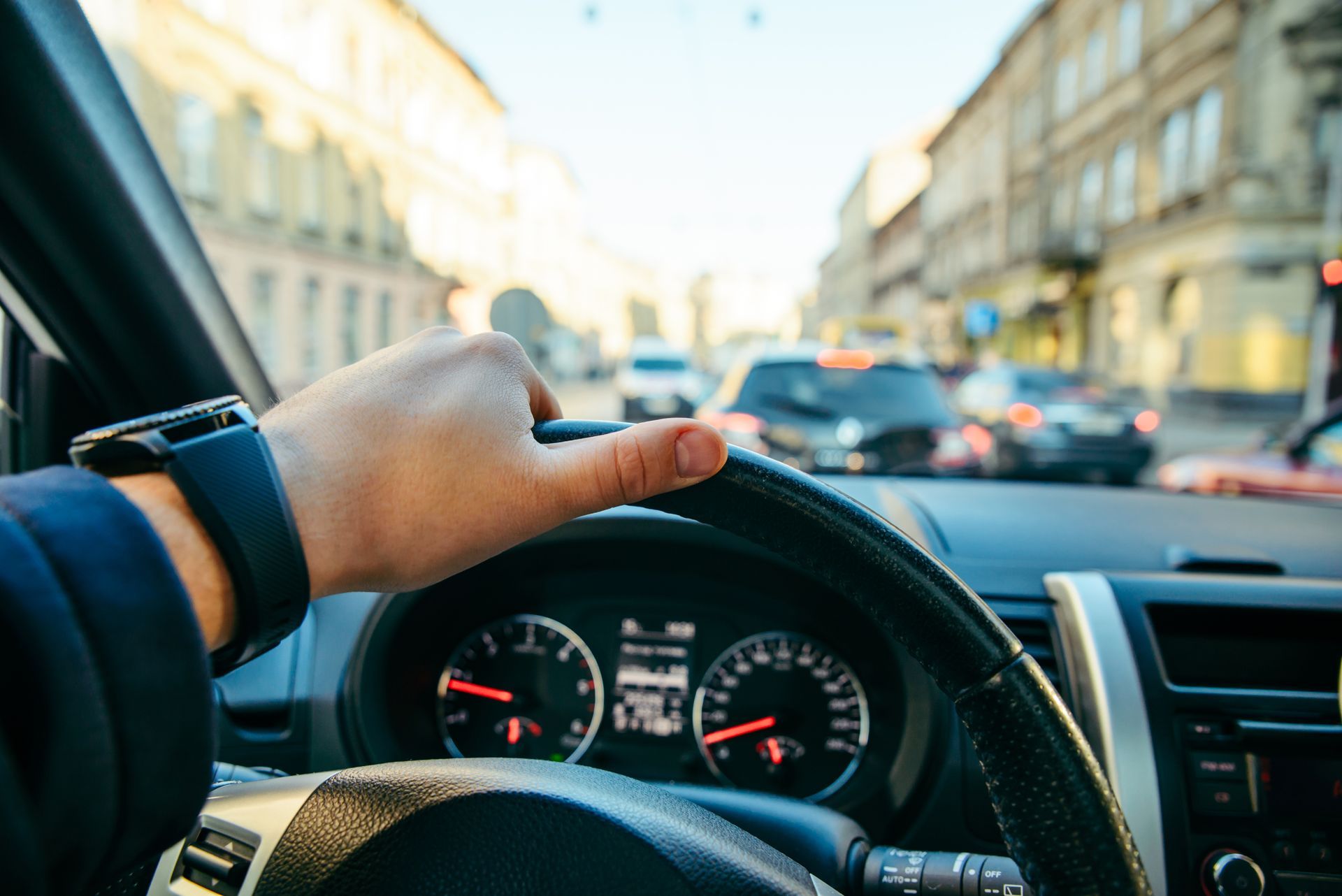 Hand gripping steering wheel driving through a city street with cars and buildings.