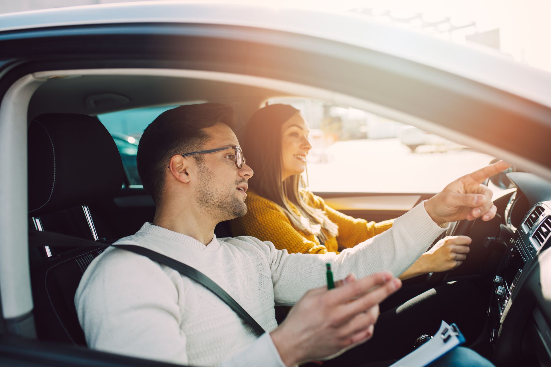 A woman driving a car with an instructor. Instructor points, smiling. Sunlight streams in. A woman driving a car with an instructor. Instructor points, smiling. Sunlight streams in.