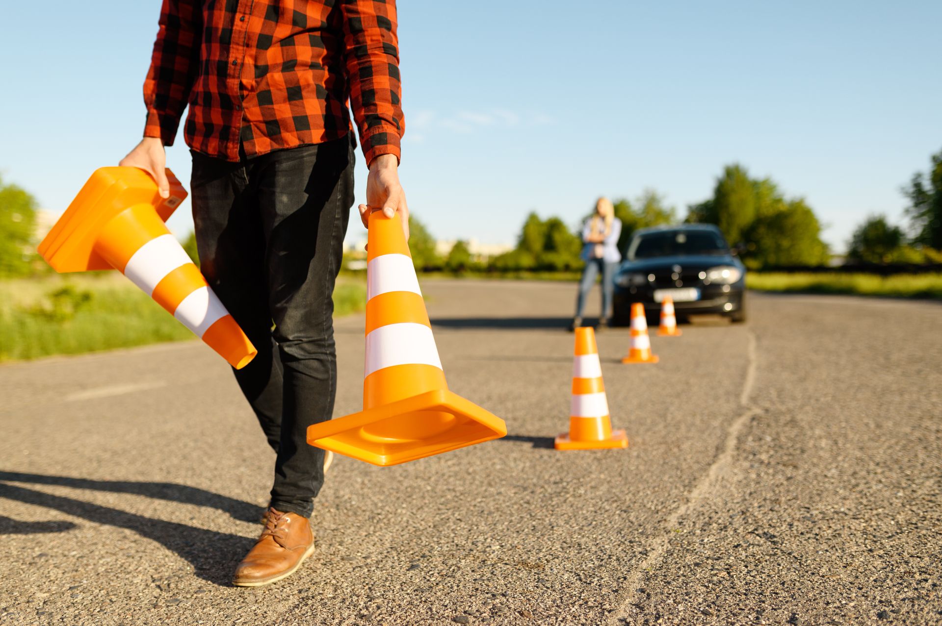 Person carrying orange traffic cones, setting up driving course. A car is in the background. Person carrying orange traffic cones, setting up driving course. A car is in the background.