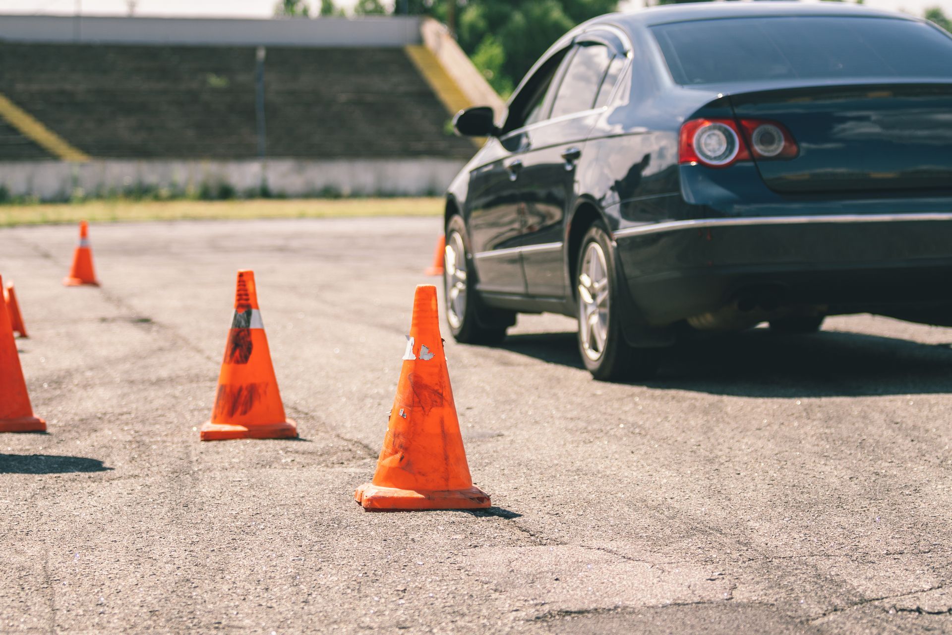 Dark blue sedan driving through orange traffic cones in a paved lot.