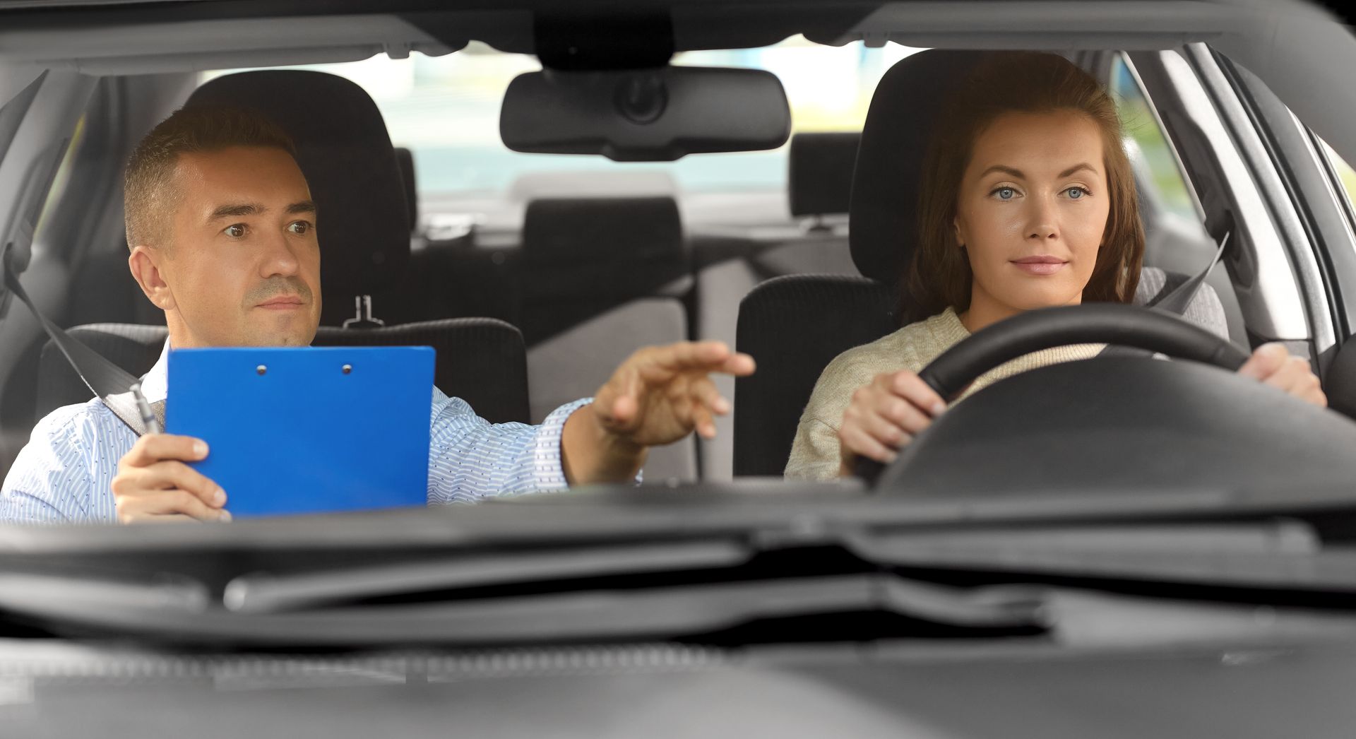 A woman driving a car with a driving instructor in the passenger seat, pointing.