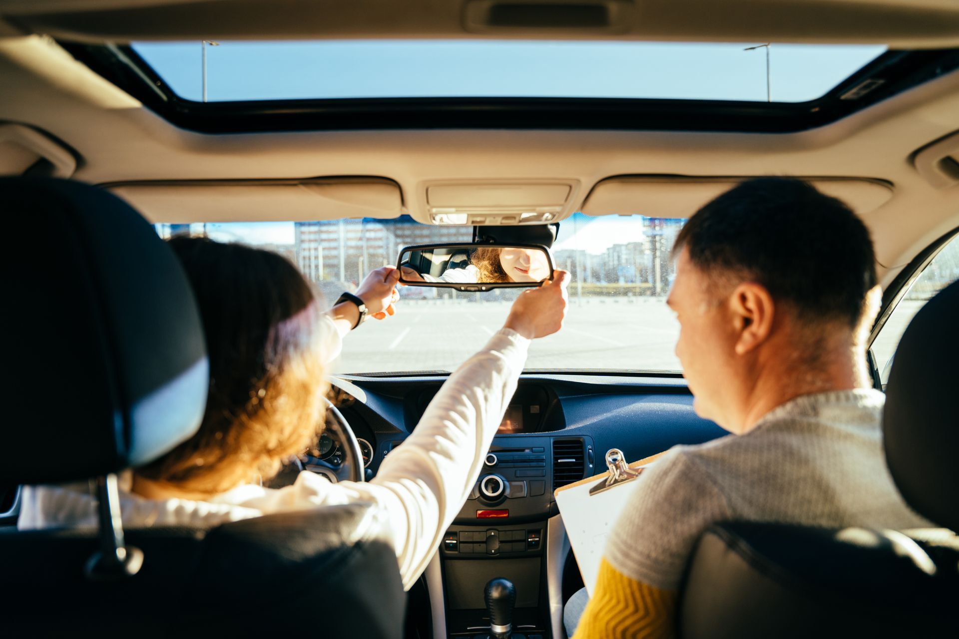 Person adjusting rearview mirror in car with instructor, sunny day.