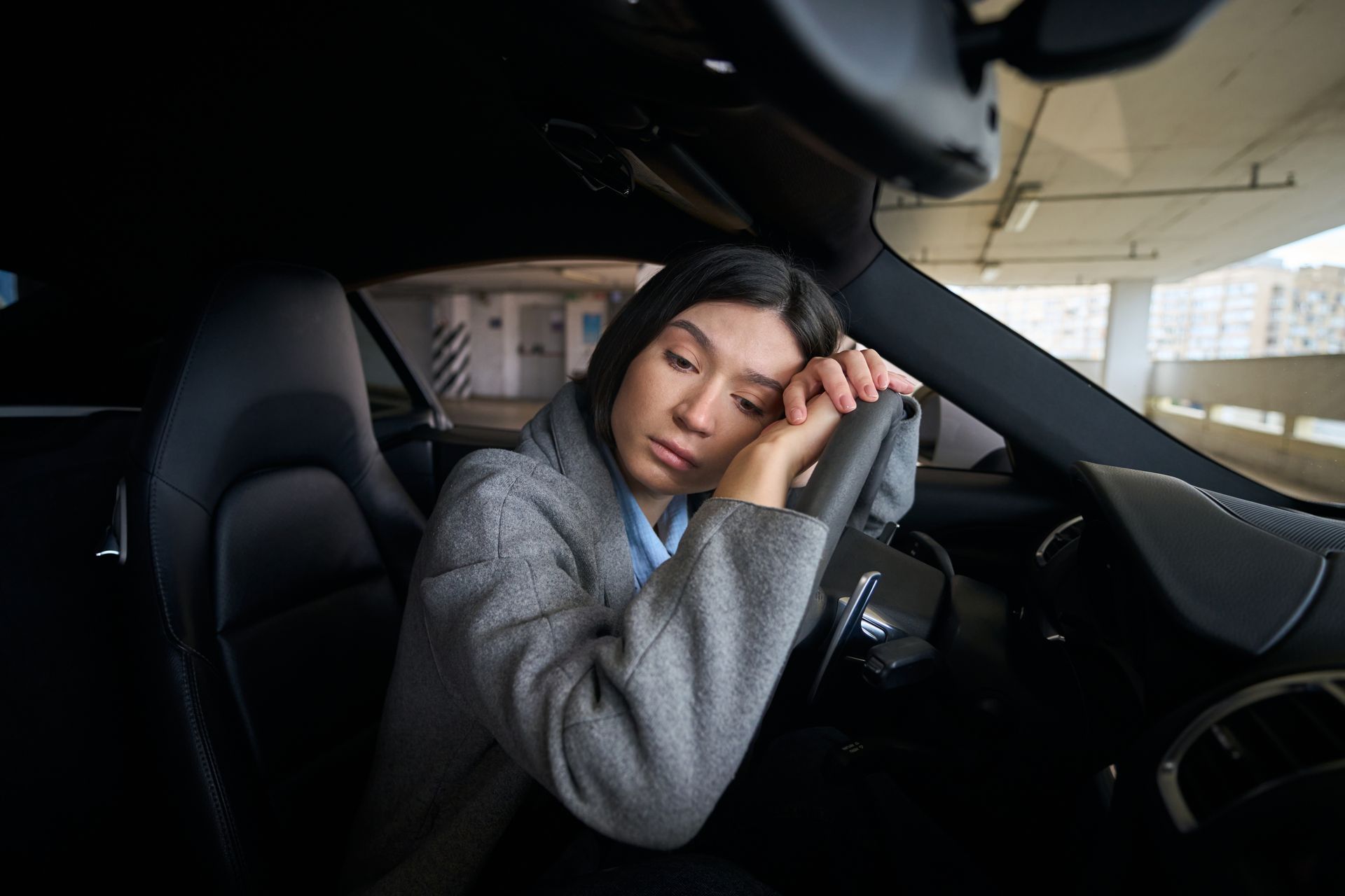 Woman resting her head on a steering wheel inside a dark car, appearing tired in a parking garage.