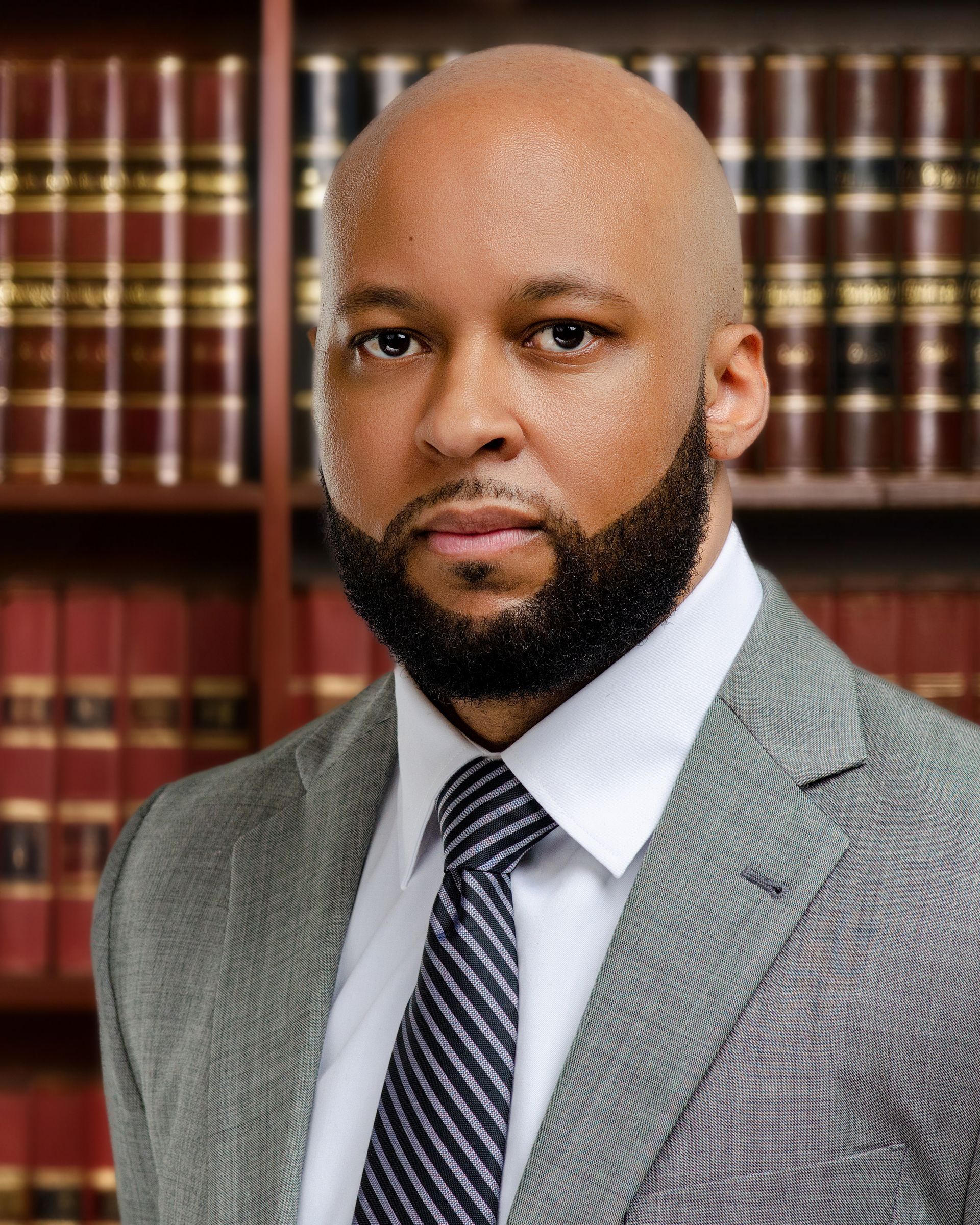 Man in a suit with a beard and bald head, standing in front of bookshelves.