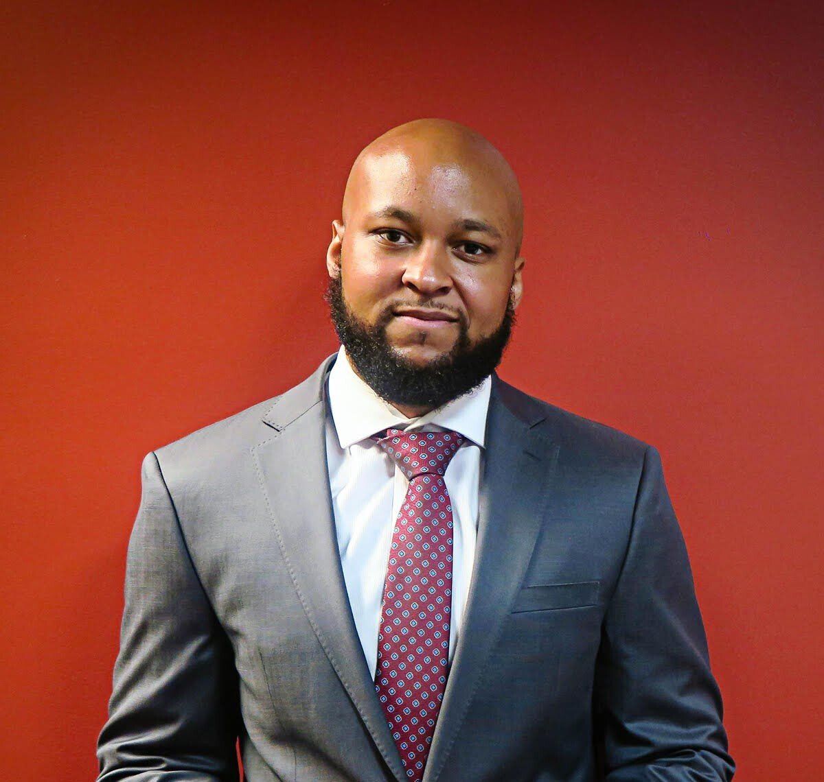 Ariel Lett with a shaved head and beard in a suit and tie against a red wall.