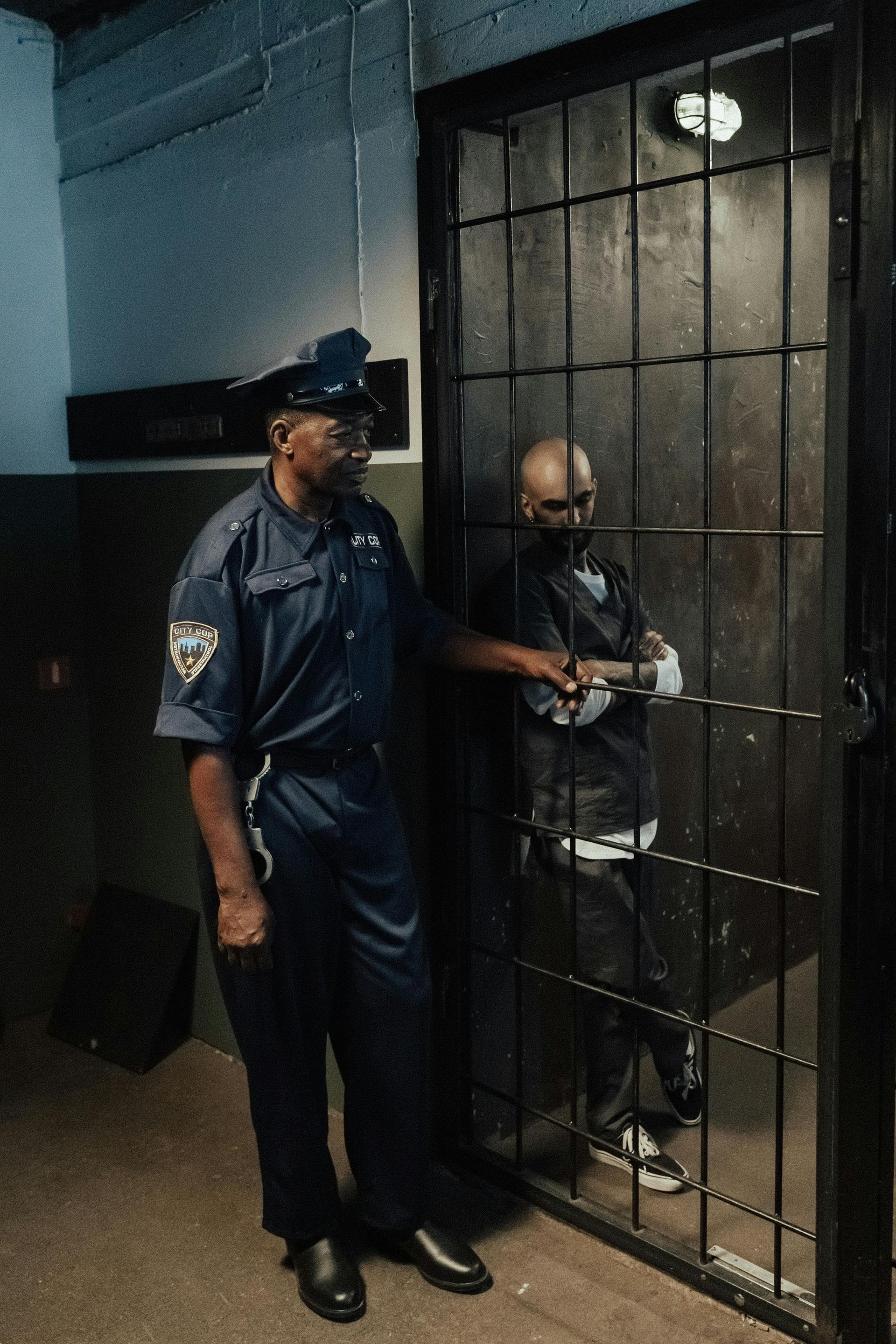 A police officer in uniform stands next to a barred cell door with a prisoner inside.