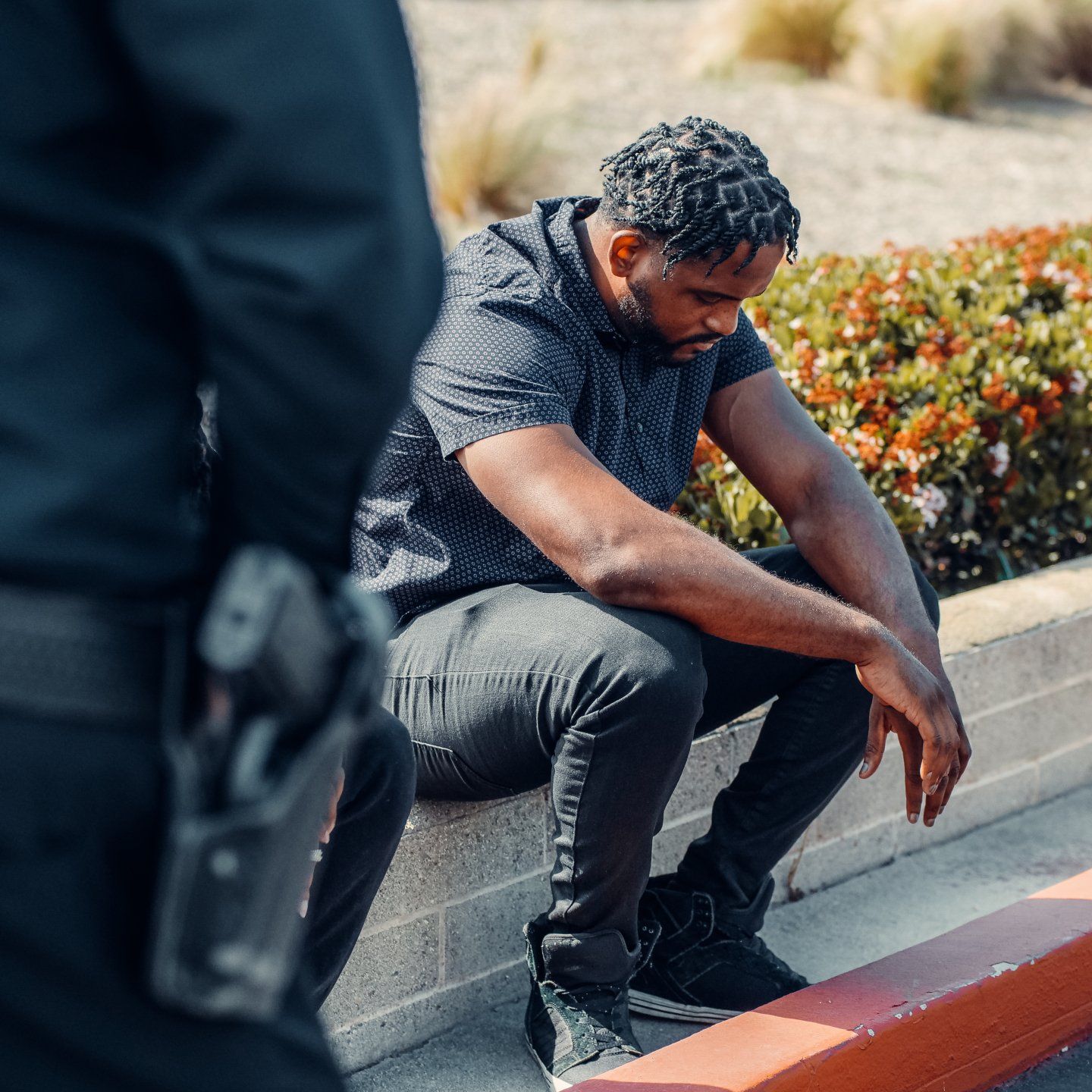 Black man seated with head down near a curb; a police officer stands nearby.