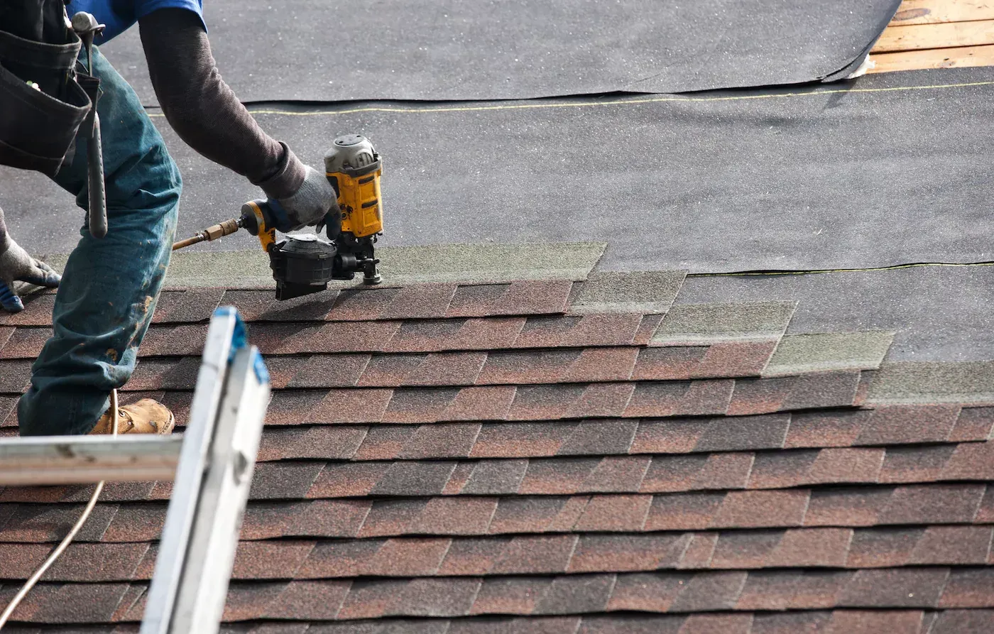 A roofer uses a yellow pneumatic nail gun to install brown asphalt shingles on a roof.