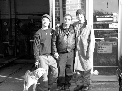 Three men and a dog stand outside a garage. One man has a rain coat. They are smiling.