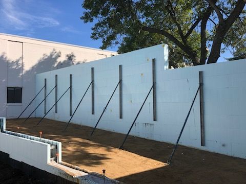 White wall with angled black supports in a dirt yard, beside a building and tree.