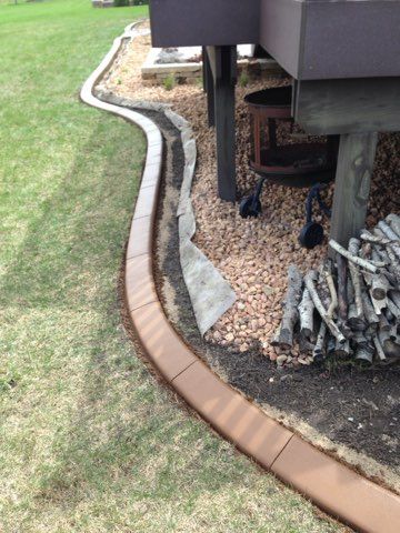 Curved brick garden edging beside a lawn and gravel area under a raised deck with stacked firewood.