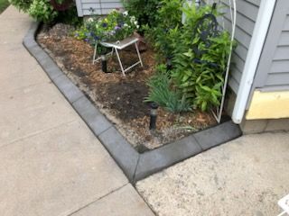Small garden bed with plants, mulch, and a folding stool beside a concrete sidewalk and house siding