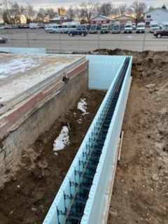 Open trench beside a concrete bridge with a blue-lined drainage channel and snowy parking lot in the background