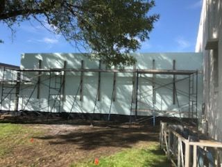 Scaffolding against a light green wall beside a white building under a tree on a sunny day