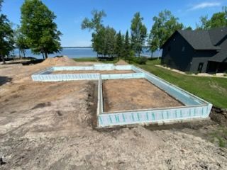 Construction site with concrete foundation footings near a lakeshore, trees, and a black house.
