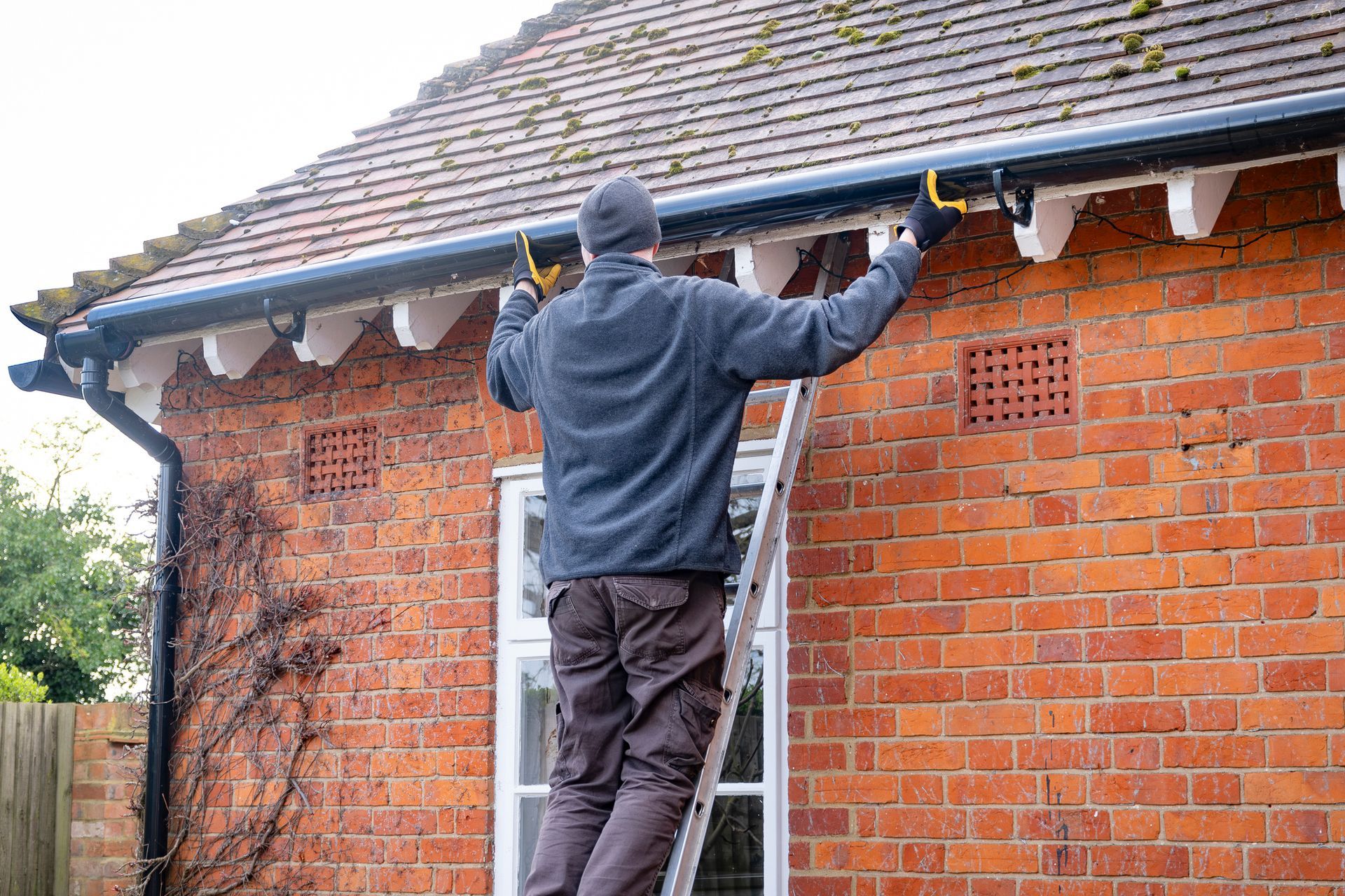 A man is standing on a ladder fixing a gutter on a brick house.