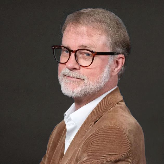 Portrait of a bearded man in glasses wearing a brown jacket and white shirt against a gray backdrop