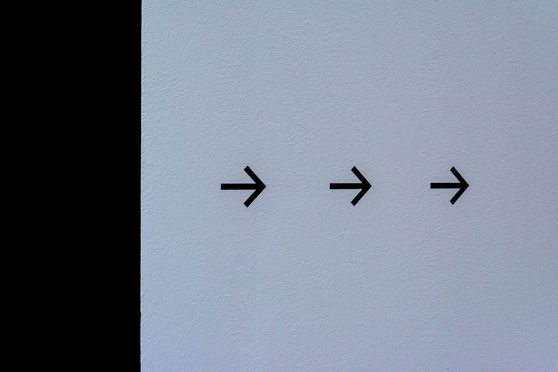 Three black right-pointing arrows on a light gray background, with a black vertical strip on the left.
