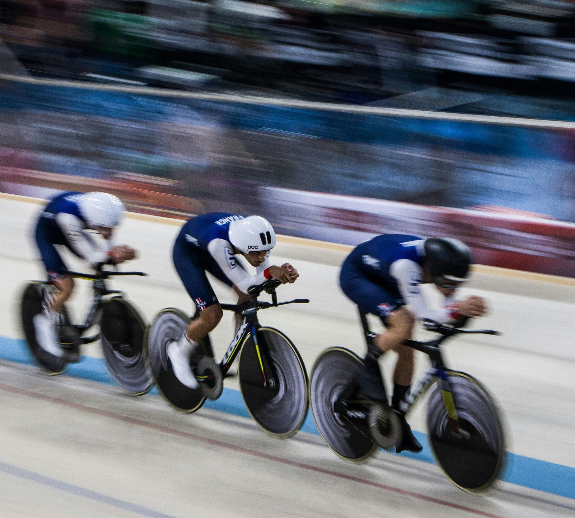 Three cyclists in blue racing bikes on a velodrome, leaning into the track in motion blur
