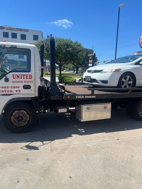 Tow truck carrying a white sedan on a sunny street near a United Auto sign