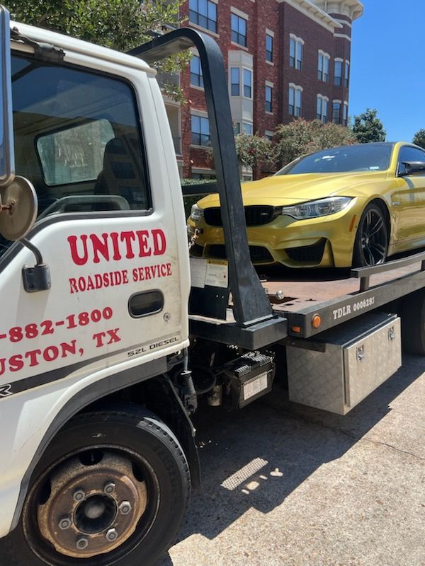 Tow truck carrying a yellow sports car on a city street, with a United Towing Service truck in the foreground