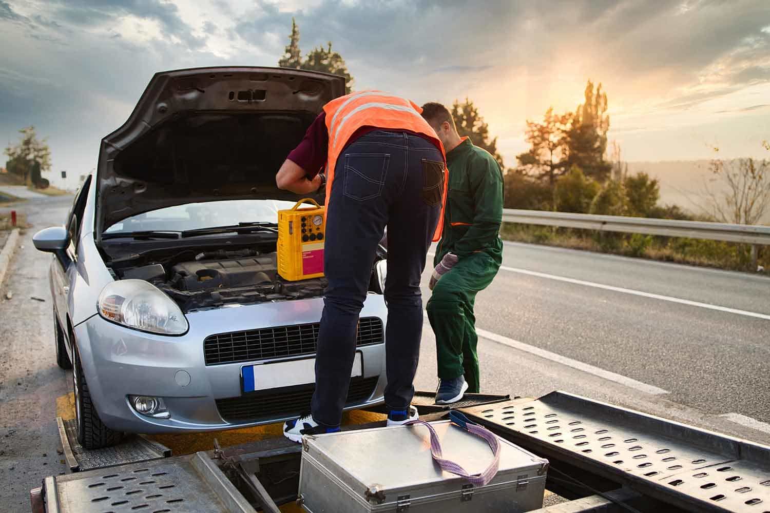 Two roadside workers inspect a silver car with open hood after a breakdown on a highway at sunset