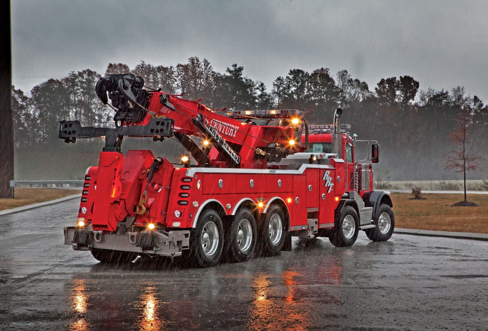Red heavy-duty tow truck with crane lights on in a rainy parking lot at dusk