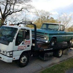 Tow truck hauling a blue vintage pickup truck on a flatbed in a roadside neighborhood.