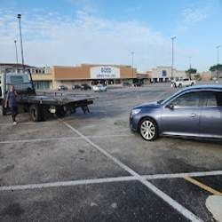 Parking lot with a tow truck and a blue sedan outside a strip mall on a sunny day