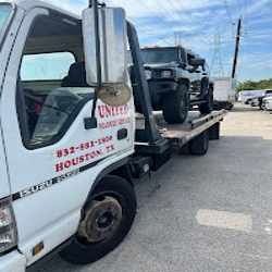 White tow truck hauling a black Jeep on a flatbed in a parking lot under a blue sky