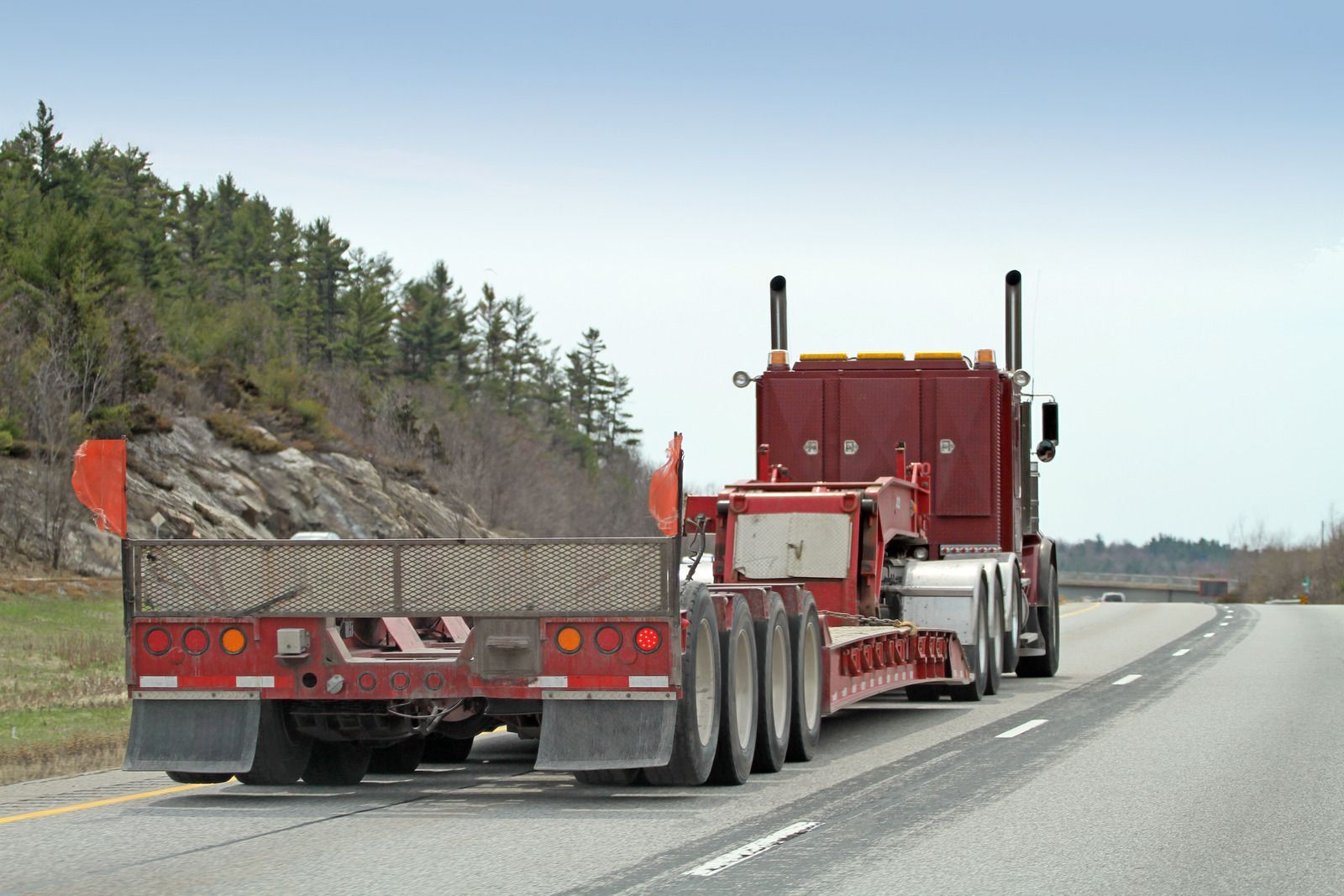Red road construction truck hauling a metal bed along a highway shoulder with trees and rocks nearby
