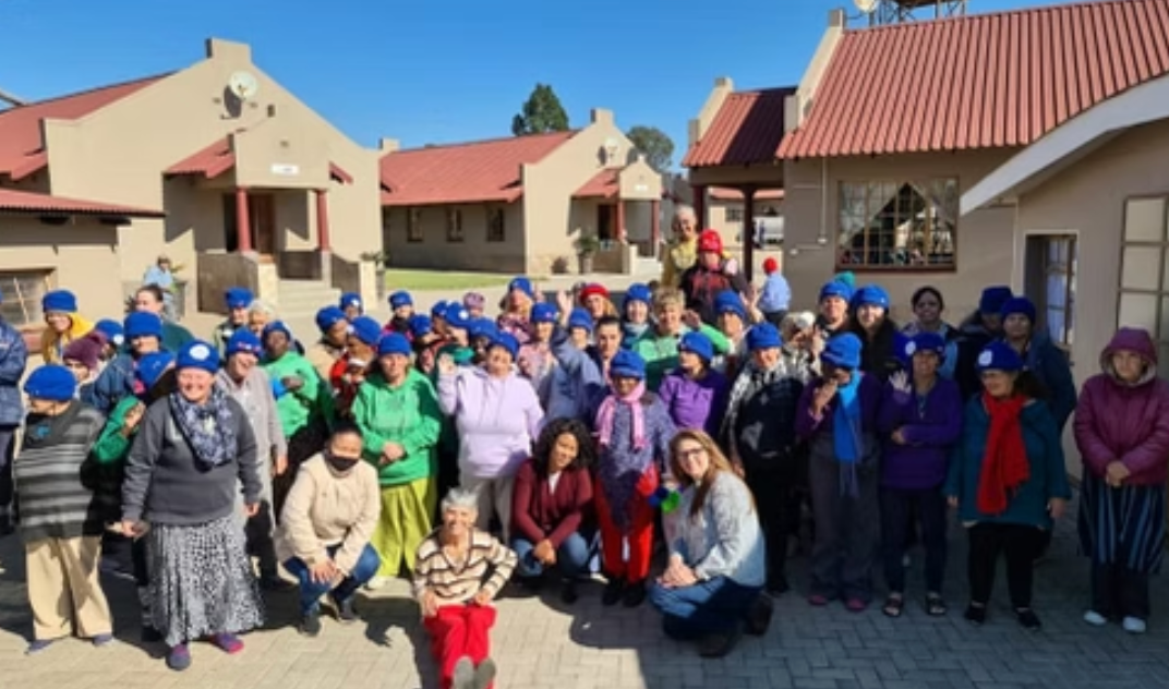 Group of people wearing blue hats pose outside beige buildings with red tile roofs.