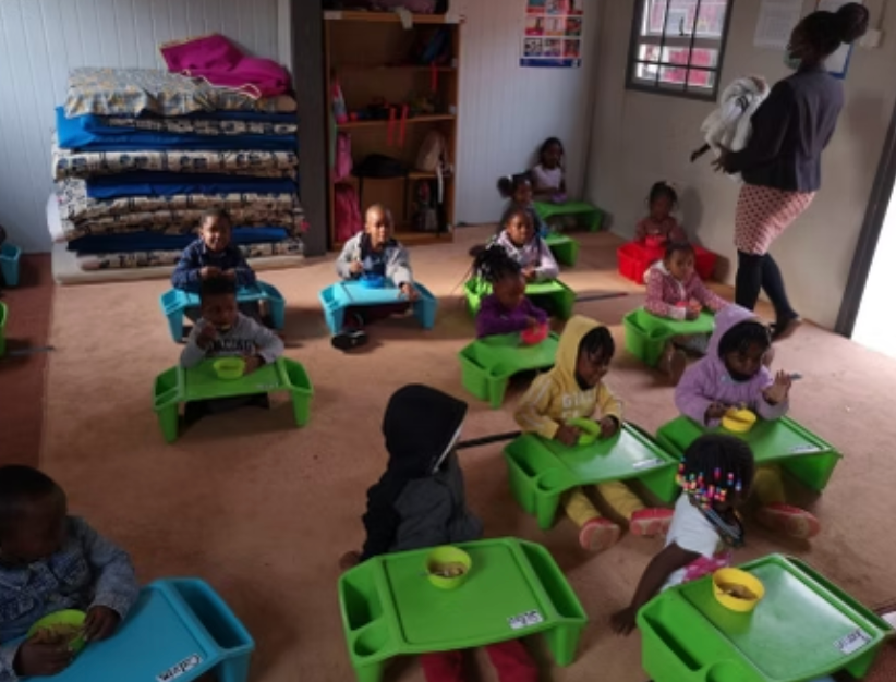 Children eating at green tables in a classroom. A woman holds a bird.