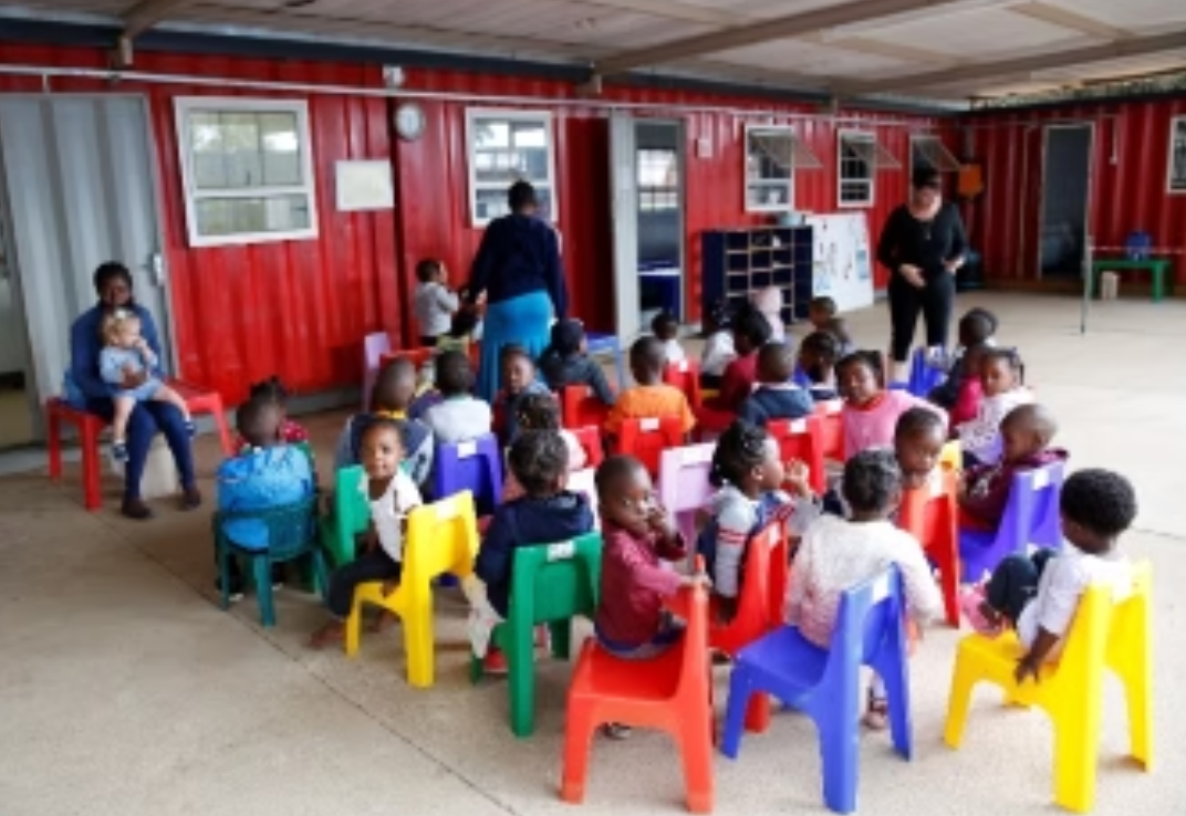 Children sitting in colorful chairs, facing teachers in a red building.