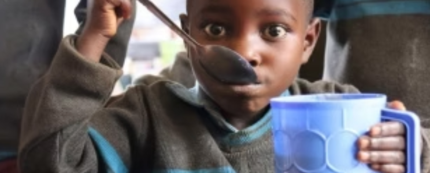 Young person holding a blue mug while eating with a spoon. Looking directly at the camera.