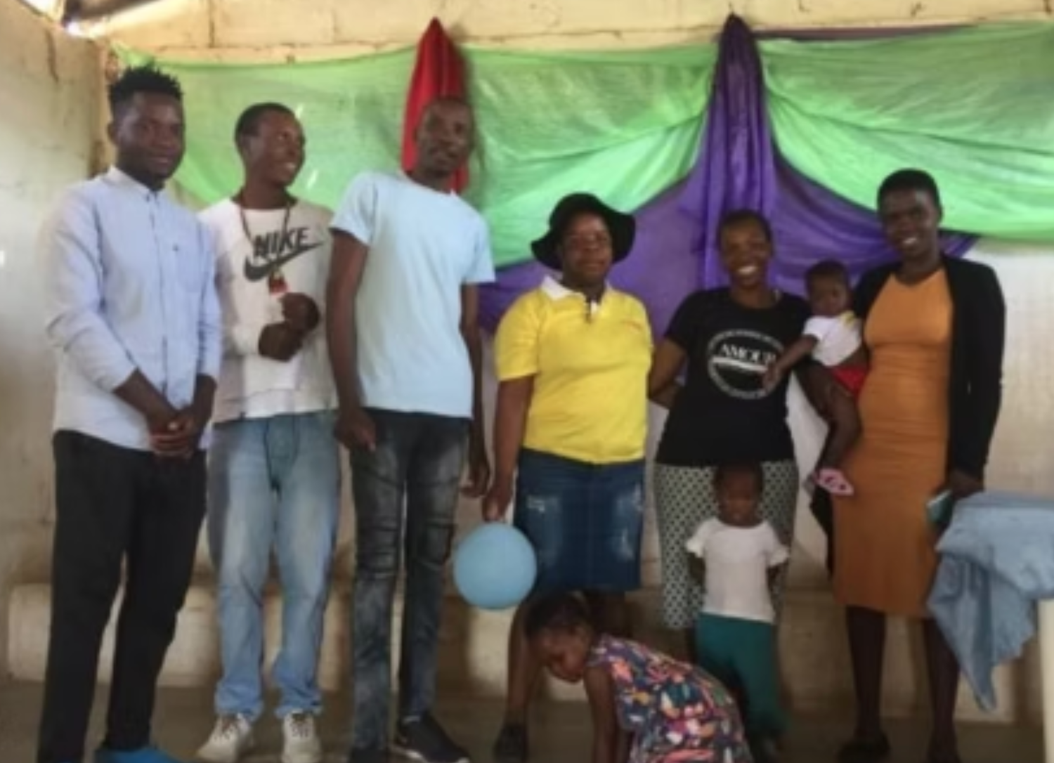 Group of people posing for a photo indoors, some holding children, with colorful fabric backdrop.