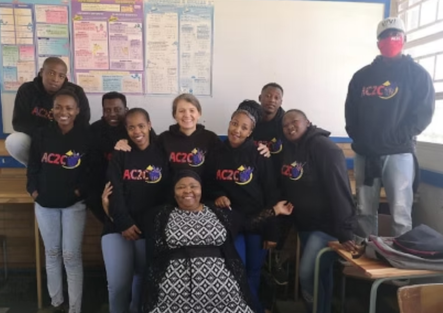 Group of people wearing black hoodies with a logo, posing in a classroom.