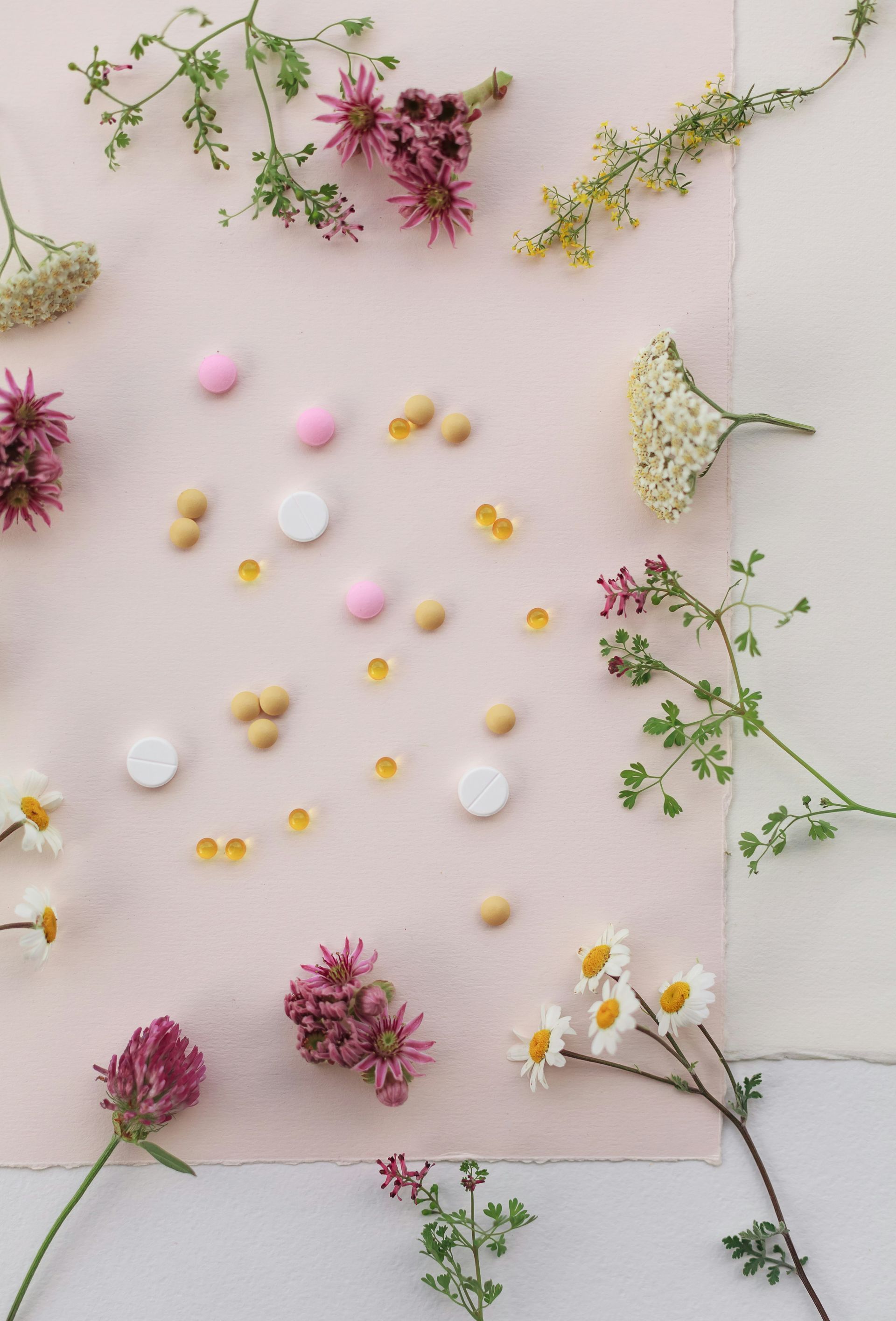 Tablets and capsules scattered on pink paper, surrounded by wildflowers and greenery.