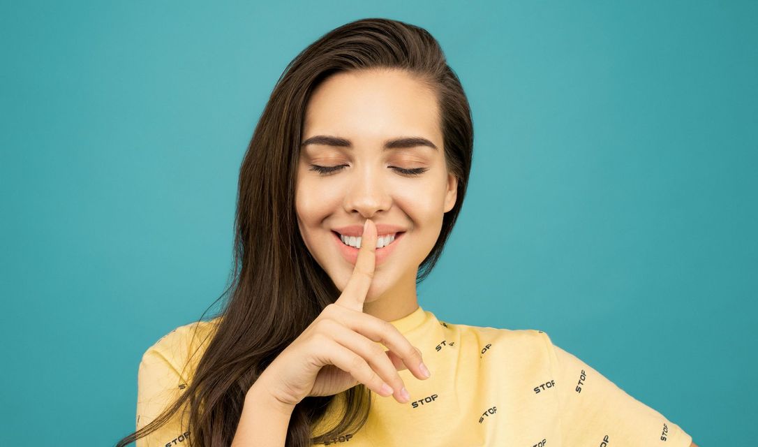 Woman with long brown hair, eyes closed, finger to lips, smiling, against a teal background.