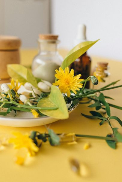 Yellow dandelions and greenery with bottles and containers on a yellow surface.